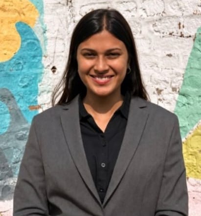 Aarushi Gupta smiling, standing in front of a brightly painted wall of marigold and teal.