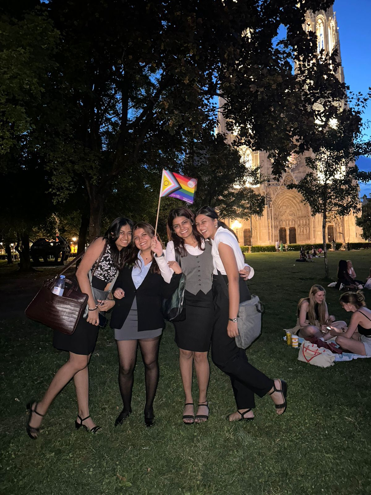 Aarushi and her teammates outdoors in Vienna at dusk, a cathedral lit behind them, holding a small pride flag.