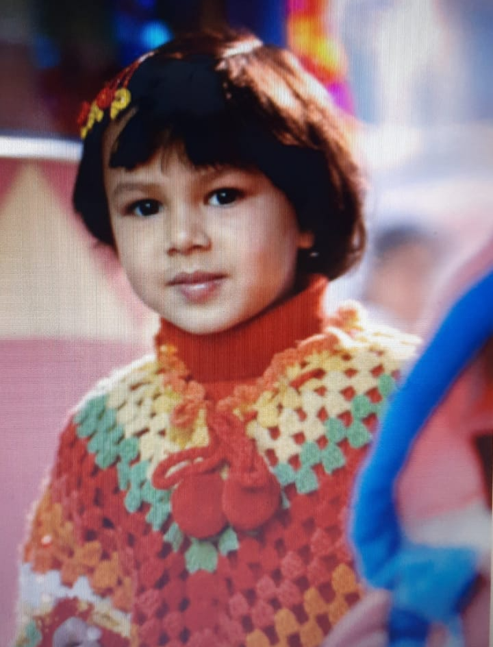 Aarushi as a young child in a colourful knitted poncho, looking into the camera.